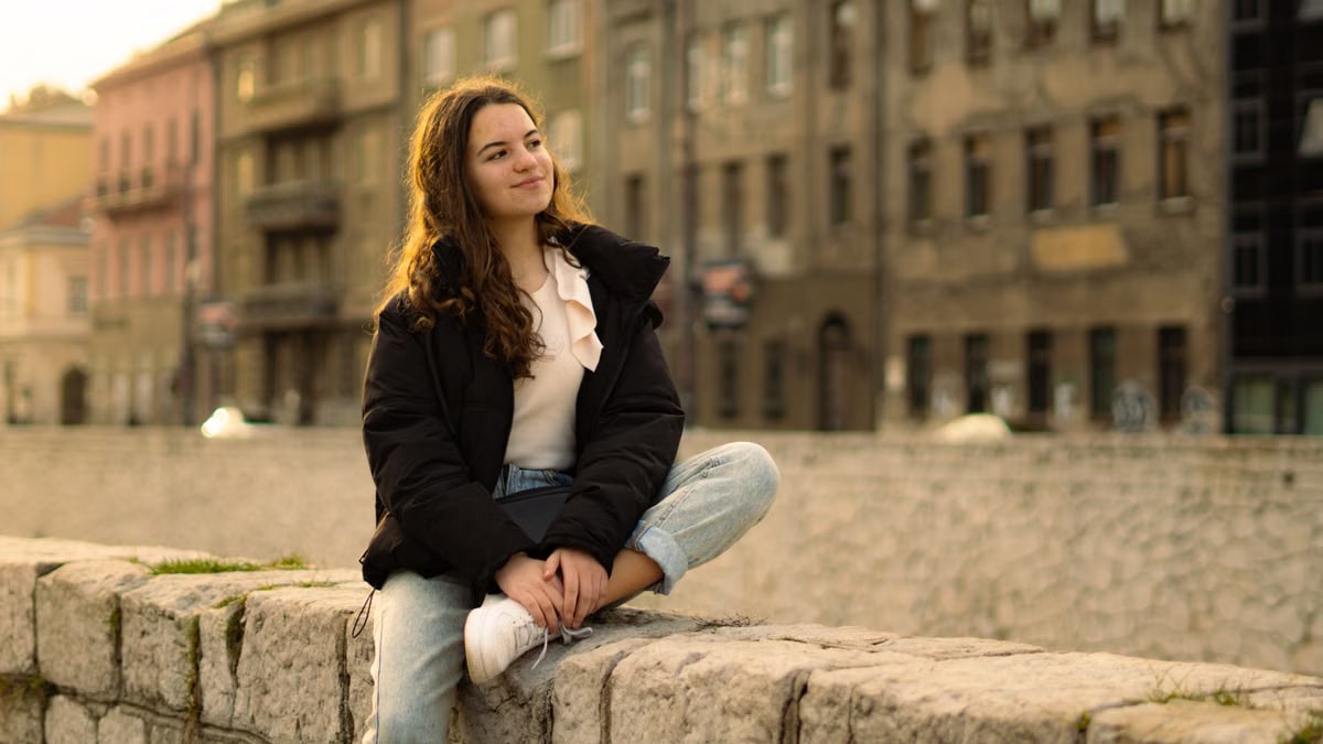 Relaxed young woman with long hair sitting on a stone wall in a cityscape.